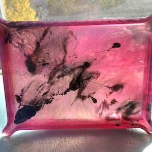 Puede incluir: Una bandeja rectangular de resina rosa con bordes curvos. El interior de la bandeja presenta una mezcla de tonos rosas y blancos, acentuados por patrones oscuros y abstractos similares a la tinta. La bandeja está diseñada con un borde ligeramente elevado y esquinas redondeadas.