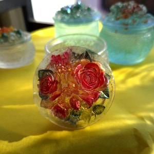 Könnte beinhalten: Ein klares, rundes Glasobjekt mit einem lebendigen roten Rosenmuster in der Mitte, umgeben von kleineren roten und gelben Blüten und grünen Blättern. Das Objekt befindet sich auf einer gelben Oberfläche, mit mehreren dekorativen Glasbehältern im Hintergrund.
