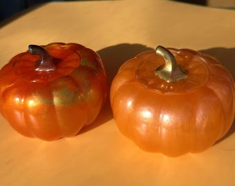 Puede incluir: Dos calabazas decorativas naranjas con detalles dorados metálicos. Cada calabaza tiene un tallo marrón oscuro y un acabado brillante. Las calabazas están sobre una superficie amarilla, ideales para la decoración otoñal.