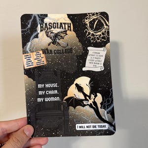 May include: A rectangular card featuring a dragon-themed design. The card includes text that reads "BASGIATH WAR COLLEGE" and "MY HOUSE. MY CHAIR. MY WOMAN." Additional text includes "I WILL NOT DIE TODAY." The design incorporates dragons, a throne, and a stormy sky.