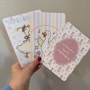 以下が含まれることがあります： 漫画のガチョウが描かれた3枚のイラストカード。1枚のカードには「OOPS！I BOI」という文字と花を持ったガチョウが描かれています。別のカードには「In a world full of serious geese be」という文字と歌うガチョウが描かれています。3枚目のカードには「don't mind me im just dilly dallying」という文字が書かれています。