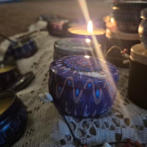May include: A collection of lit candles in patterned tins, with blue and purple designs, are arranged on a lace cloth. The flames cast a warm light, creating a cosy atmosphere. The candles are in various sizes, suggesting a decorative theme.