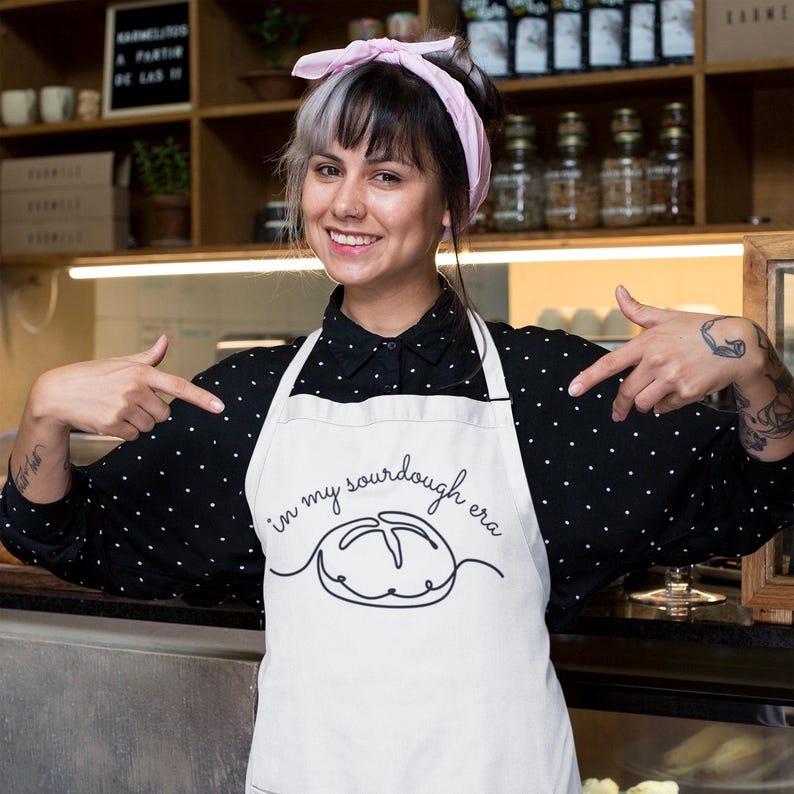 May include: A white apron with black text that reads "in my sourdough era" and a line drawing of a loaf of bread. The person wearing the apron is smiling and pointing at the apron.
