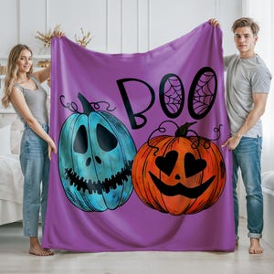 May include: A purple Halloween blanket featuring the word "BOO" with spiderwebs and two cartoon pumpkins. One pumpkin is blue with a carved face, and the other is orange with heart-shaped eyes. The blanket is held up by two people.