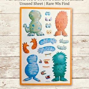 May include: A sheet of vintage AGC monster stickers. The stickers feature various colorful monster designs, including blue, orange, and green creatures, along with eyes, claws, and teeth. The sheet is bordered in orange and includes the text "VINTAGE AGC MONSTER STICKERS".