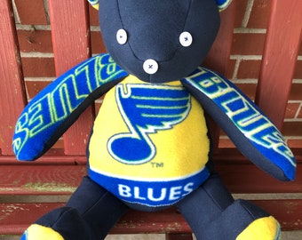 May include: A blue and yellow teddy bear wearing a St. Louis Blues jersey. The bear has button eyes and a blue nose. The jersey has a blue note on the front with the word "Blues" written below it.
