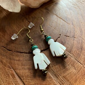 May include: A pair of dangle earrings featuring white carved shell bird pendants, small round turquoise beads, and antique brass-toned metal accents. The earrings are suspended from brass-toned ear wires with clear silicone backings.