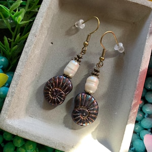 May include: A pair of handcrafted earrings featuring iridescent, dark purple ammonite-shaped beads. Each earring has a white pearl and antique brass-colored accents. The earrings are displayed in a gray, rectangular dish.