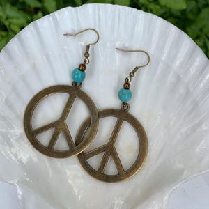 May include: A pair of bronze-colored peace sign earrings with turquoise beads. The earrings are displayed on a white seashell against a backdrop of green foliage. The earrings have a vintage, bohemian style.