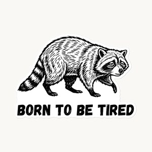 以下が含まれることがあります： 疲れた表情で歩くアライグマの白黒イラスト。アライグマの下には「BORN TO BE TIRED」というテキストがあります。アライグマは縞模様の尻尾を持ち、四足歩行で歩いています。