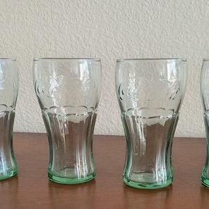 May include: Four clear drinking glasses with a green base. The glasses have a curved shape and a decorative embossed design. They are arranged in a row on a brown surface.