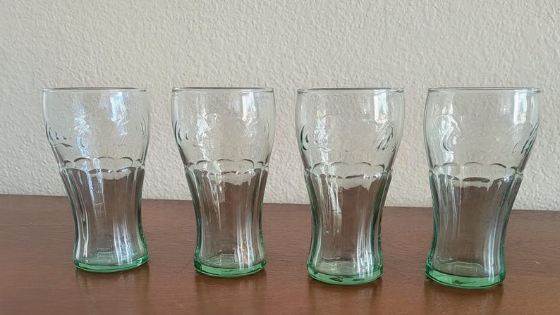May include: Four clear drinking glasses with a green base. The glasses have a curved shape and a decorative embossed design. They are arranged in a row on a brown surface.