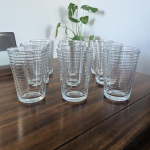 May include: A set of six clear glass tumblers, each featuring a series of horizontal etched rings encircling the exterior. The glasses have a slightly tapered cylindrical shape with a smooth, flat base and a clear, transparent finish throughout.