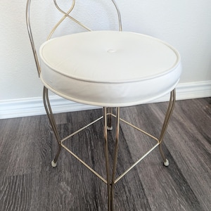 May include: A vanity stool featuring a round, white upholstered seat with a central button tuft. The frame is constructed from thin, polished gold-tone metal tubing, forming four legs and a curved backrest with a decorative looped design.
