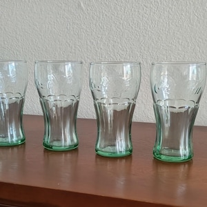 May include: Four clear Coca-Cola glasses with a green base. The glasses have a classic shape and are arranged on a dark brown surface. The Coca-Cola logo is visible on each glass. The glasses are approximately 15 cm tall.