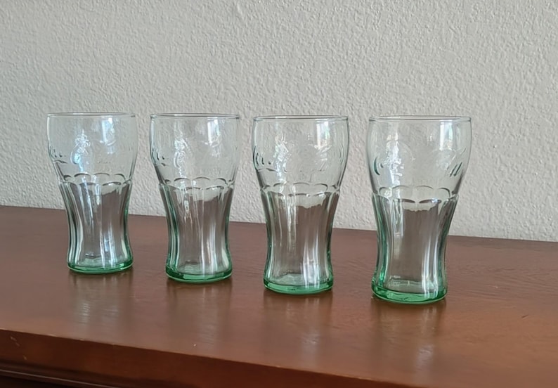 May include: Four clear Coca-Cola glasses with a green base. The glasses have a classic shape and are arranged on a dark brown surface. The Coca-Cola logo is visible on each glass. The glasses are approximately 15 cm tall.