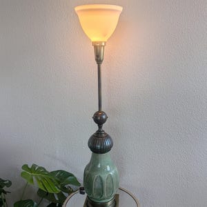 May include: An antique floor lamp with a green ceramic base and a brass stem. The lamp has a cream-colored, cone-shaped shade. The lamp is on a mirrored table with a plant in the foreground.
