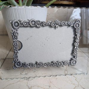 May include: A clear glass photo frame with a decorative border of metal sunflowers. The frame has a scalloped edge and is designed to hold a rectangular photo. The metal sunflowers are detailed and add a touch of elegance.