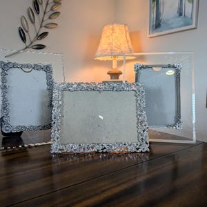 May include: Three rectangular picture frames with silver-toned, textured floral borders. Two frames are oriented vertically with clear acrylic edges, while the central frame is oriented horizontally with a dense, embossed metallic floral pattern.