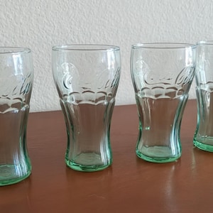 May include: Four clear Coca-Cola glasses with a green tint at the base. The glasses have a classic, curved shape and the Coca-Cola logo is etched on the front. They are arranged on a brown surface.