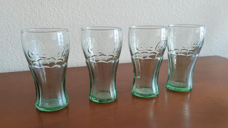 May include: Four clear Coca-Cola glasses with a green tint at the base. The glasses have a classic, curved shape and the Coca-Cola logo is etched on the front. They are arranged on a brown surface.