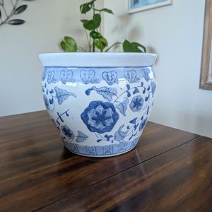 May include: A round ceramic planter featuring a white base with blue floral and vine patterns. The rim is solid white, while the upper and lower sections are decorated with a repeating blue border design. The planter has a smooth, glossy finish.
