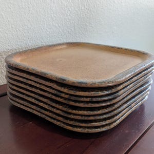 May include: A stack of square ceramic plates with rounded corners. The plates have a warm brown color with a textured, dark brown rim. The plates are stacked on top of each other, showcasing their unique design and shape.