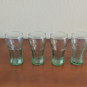 May include: Four clear Coca-Cola glasses with a green tinge, arranged in a row on a brown wooden surface. The glasses have a classic, curved shape and are empty. The background is a plain, off-white wall.
