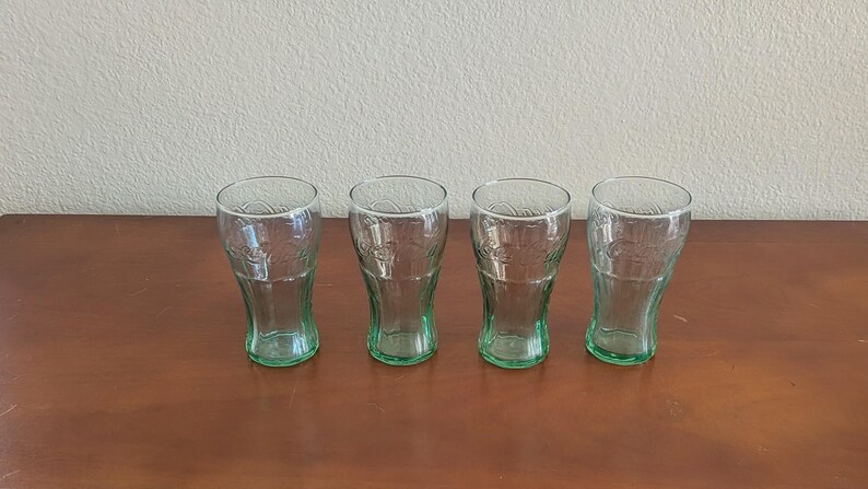 May include: Four clear Coca-Cola glasses with a green tinge, arranged in a row on a brown wooden surface. The glasses have a classic, curved shape and are empty. The background is a plain, off-white wall.