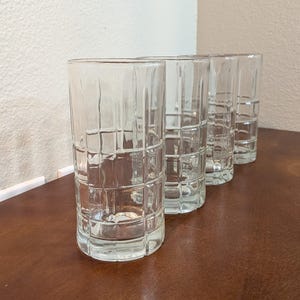 May include: Four clear, textured highball glasses with a grid pattern. The glasses are cylindrical with a thick base and are arranged in a row on a brown surface. The glasses are made of clear glass.