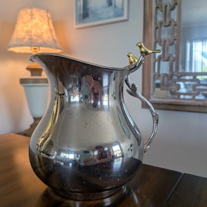 May include: A polished silver-toned metal pitcher featuring a curved handle shaped like a branch. Two small, gold-toned bird figures are perched on the top of the handle where it meets the rim of the pitcher. The surface is reflective and smooth.