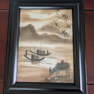 May include: Framed artwork depicting a serene landscape with boats on a river, mountains in the background, and a person riding a water buffalo. The artwork is in sepia tones and framed in black.