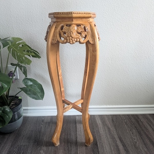 May include: A tall, light brown wooden plant stand featuring an octagonal top with a beaded edge. The apron is decorated with carved grape and leaf motifs. It has four curved legs connected by a central cross-shaped stretcher near the base.