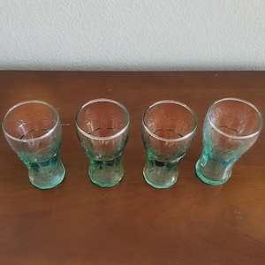 May include: Four vintage Coca-Cola glasses. The glasses are made of clear glass with a green tint and a silver rim. The glasses have a unique shape and are arranged on a brown wooden surface.