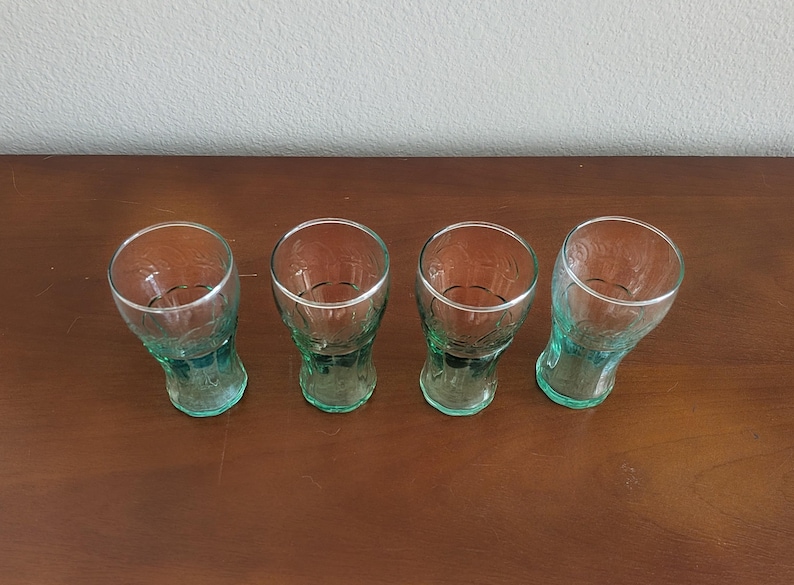 May include: Four vintage Coca-Cola glasses. The glasses are made of clear glass with a green tint and a silver rim. The glasses have a unique shape and are arranged on a brown wooden surface.