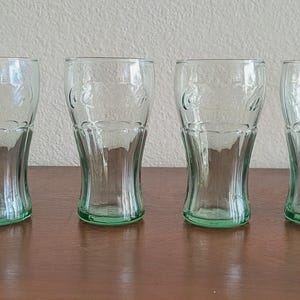May include: Four clear, green-tinted Coca-Cola glasses. Each glass has a curved shape and the Coca-Cola logo embossed on the front. The glasses are displayed on a brown surface.
