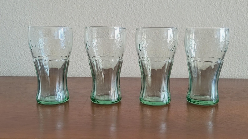 May include: Four clear, green-tinted Coca-Cola glasses. Each glass has a curved shape and the Coca-Cola logo embossed on the front. The glasses are displayed on a brown surface.