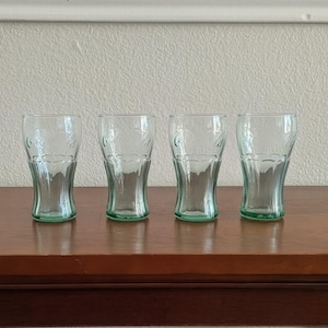 May include: Four clear, green-tinted Coca-Cola glasses are arranged on a dark wooden surface. The glasses have a classic, curved shape and are etched with the Coca-Cola logo. The glasses are empty and ready to be filled.