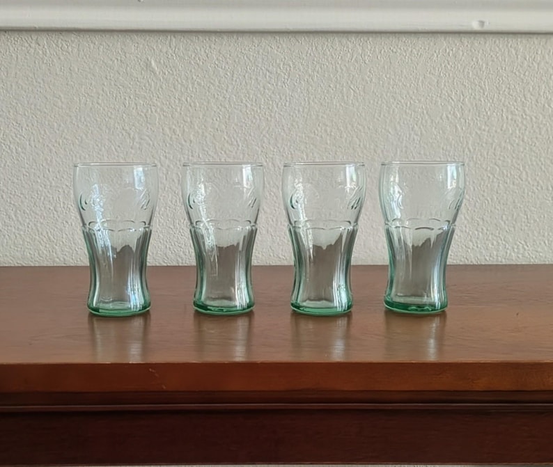 May include: Four clear, green-tinted Coca-Cola glasses are arranged on a dark wooden surface. The glasses have a classic, curved shape and are etched with the Coca-Cola logo. The glasses are empty and ready to be filled.