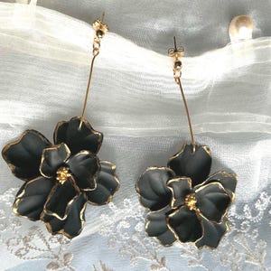 Puede incluir: Un par de pendientes de flores negras con ribetes dorados. Los pendientes tienen un alambre dorado largo y delgado y una pequeña cuenta dorada en la parte superior. Los pétalos de la flor son negros con bordes dorados y un centro dorado.