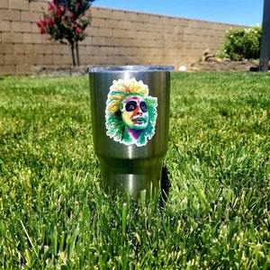 May include: A stainless steel tumbler with a colorful sticker of a character from the movie "Beetlejuice". The sticker features a green, yellow, and purple character with a surprised expression.