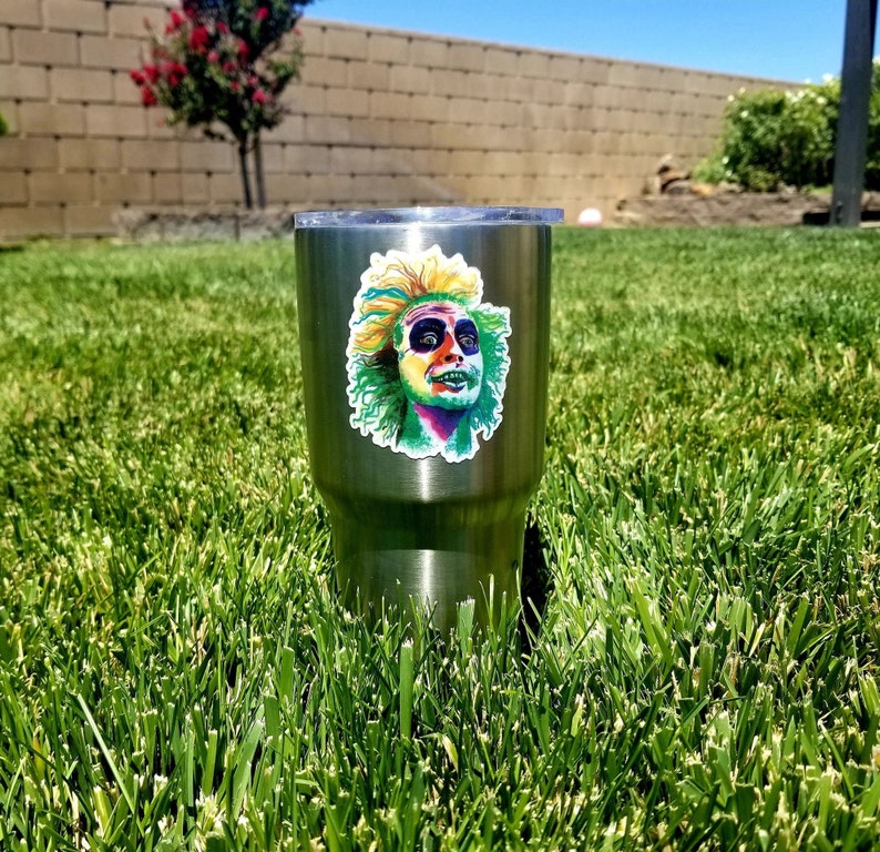 May include: A stainless steel tumbler with a colorful sticker of a character from the movie "Beetlejuice". The sticker features a green, yellow, and purple character with a surprised expression.