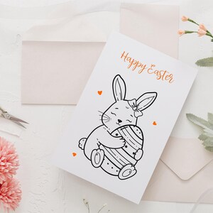 May include: White greeting card with the text "Happy Easter" in orange script. The card features a black and white illustration of a bunny hugging a decorated Easter egg. Small orange hearts are scattered around the card.