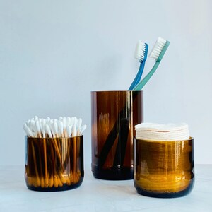 May include: Three amber glass containers of different sizes. One holds cotton buds, another has two toothbrushes, and the third contains cotton pads. The cylindrical containers are on a white surface.