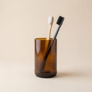 Può includere: Un bicchiere di vetro marrone con due spazzolini da denti. Uno spazzolino ha una testina con setole bianche, l'altro con setole nere. Gli spazzolini sono in legno e di colore naturale.