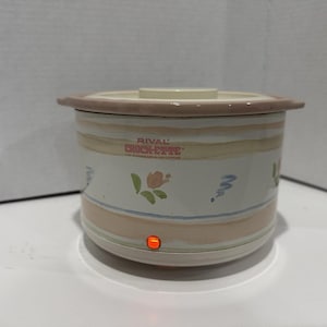 May include: A Rival Crock-ette slow cooker with a cream-colored lid and a light pink rim. The white body features a floral and swirl design with horizontal stripes in peach and tan. The power indicator light is illuminated.