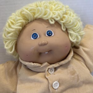 May include: A vintage doll with a soft, tan face, blue eyes, and a tuft of yellow yarn hair. The doll is wearing a tan corduroy outfit with white buttons. The doll has a small, open mouth with two teeth.