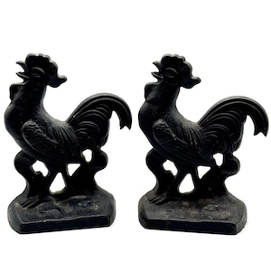 May include: Two black cast iron rooster figurines. Each rooster is standing on a base with a detailed design. The roosters have detailed feathers and a proud stance. The background is white.