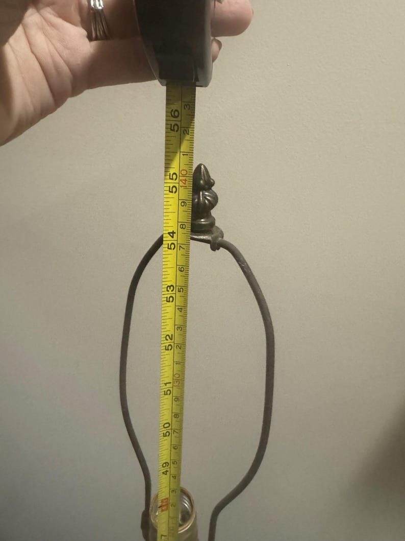 May include: A close-up of a lamp base with a metal harp and a decorative finial. The base is being measured with a tape measure, which reads 56 inches.
