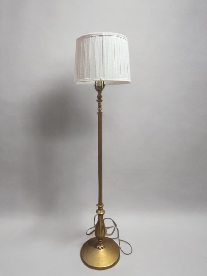 May include: A gold floor lamp with a white pleated lampshade. The lamp has a simple design with a round base and a tall, slender stem.